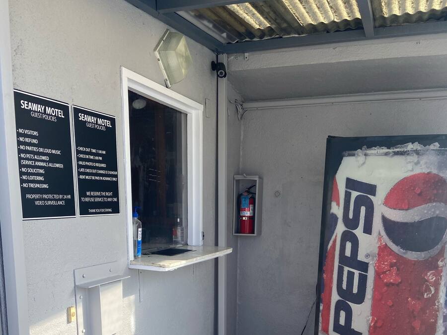 A corner of a building with a white wall, a window, and a door. There are two signs on the wall listing guest policies for a motel. A fire extinguisher is mounted on the wall, and a Pepsi vending machine is visible.