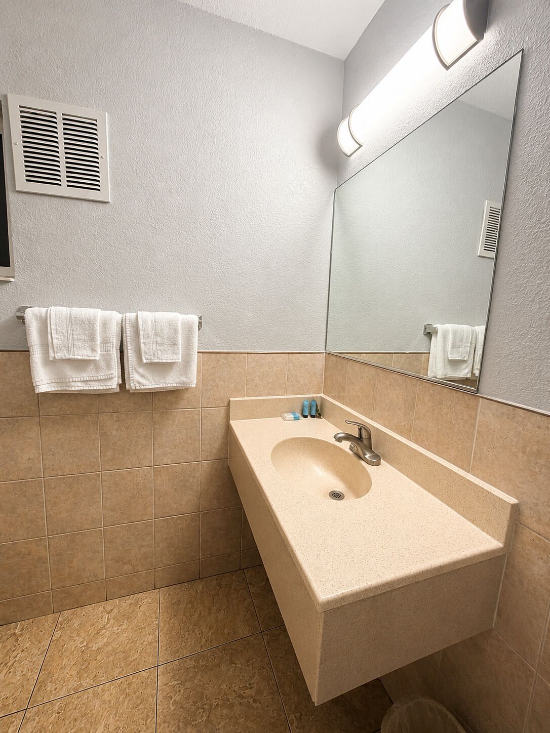 A bathroom with a beige sink, two white towels, a mirror, and a ventilation vent.