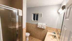 A bathroom with a glass shower enclosure, a toilet, a sink with a mirror, and a window with a vent. - Seaway Motel (Los Angeles)