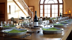 Uma longa mesa de jantar posta com taças de vinho, pratos e uma garrafa de vinho, num espaço com escadas e janelas amplas.