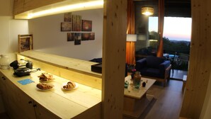 A wooden counter with pastries and a coffee maker, a living area with a couch and a view of the sunset, and a dining table with a vase and a lamp. - Heavenly Duplex with 2 gardens at Redrock Faqra (faqra)