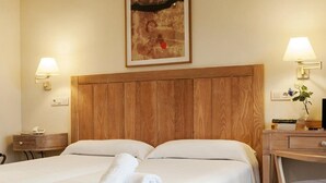 Quarto de hotel com cabeceira de madeira, cama com roupa de cama branca, escrivaninha com cadeira e duas luminárias fixadas na parede.
