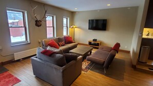 A spacious living room with a grey sofa, a brown armchair, and a coffee table. There are two windows with curtains, a mounted television, and a wall-mounted deer head. - Modern Downtown Loft in Heart of Silverton (Silverton)