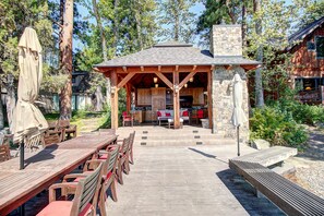 An outdoor dining area with wooden tables and chairs, a stone building with a chimney, and a view of a forested area.