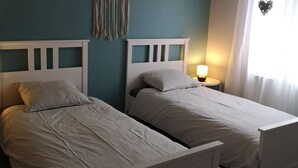 Une chambre avec deux lits simples, une décoration en macramé suspendue, un luminaire blanc au plafond et une fenêtre avec des rideaux légers.