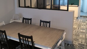 Un espace repas avec une grande table et des chaises, une vue sur la cuisine à travers une fenêtre et des carreaux de sol à motifs.