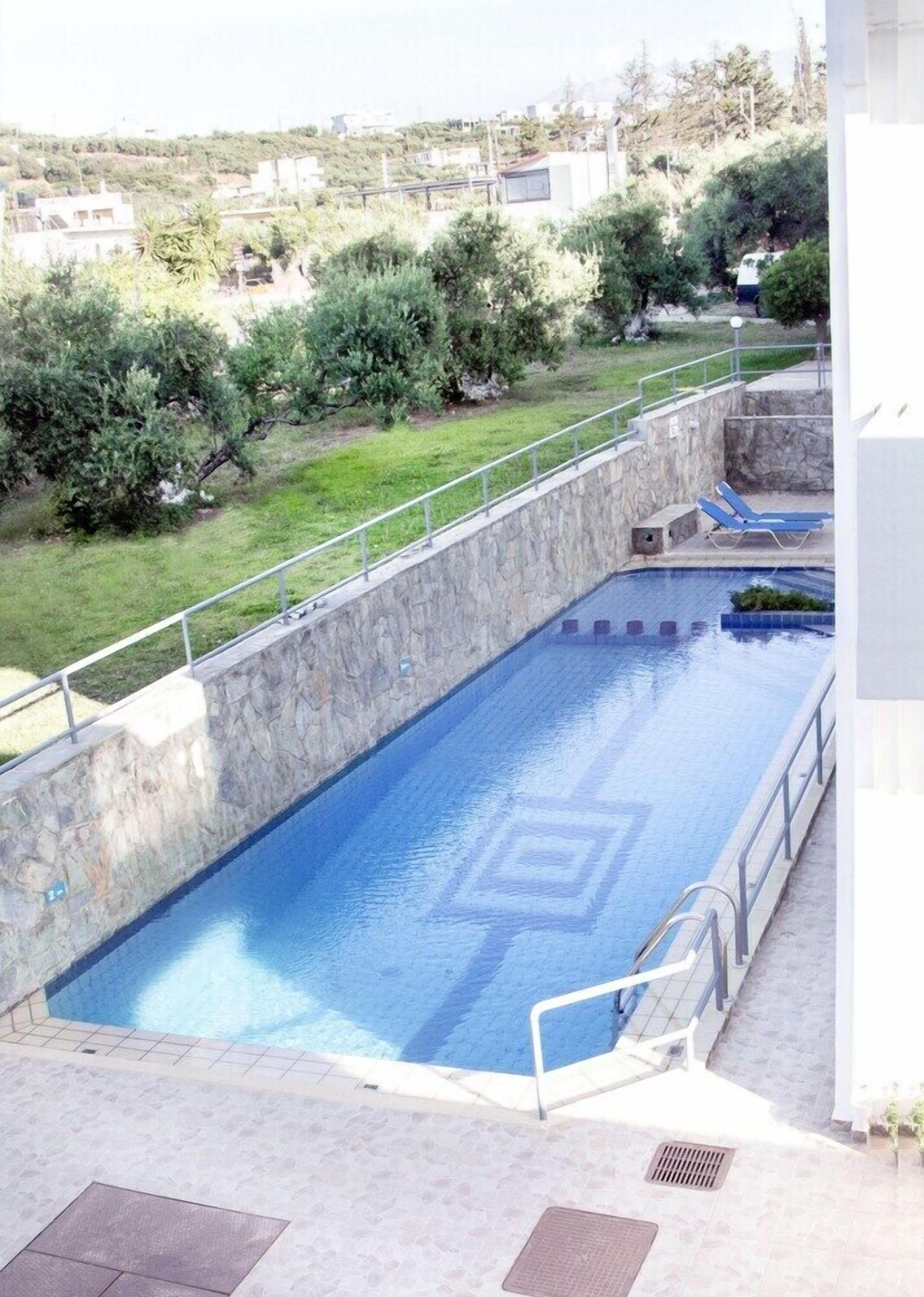 En swimmingpool med en mur av sten och ett metallräcke. Det finns solstolar och en gräsyta med träd och buskar i bakgrunden.