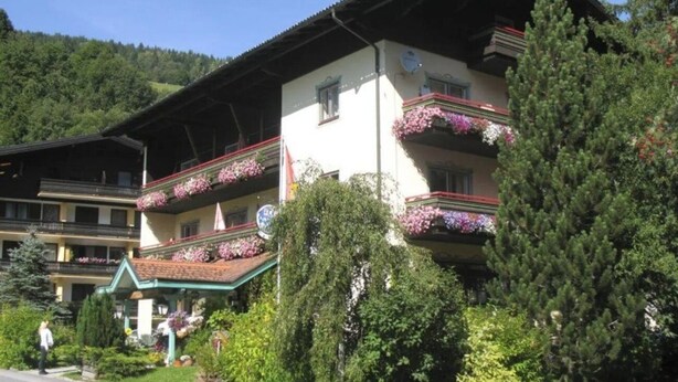 Un edifici de diversos pisos amb balcons decorats amb flors, envoltat de vegetació i un paisatge de muntanyes al fons.