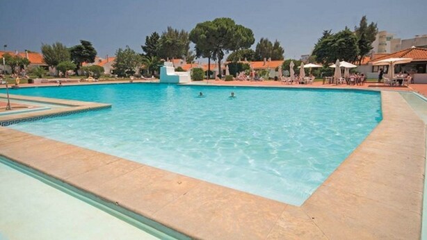 A large outdoor swimming pool with clear blue water, surrounded by lounge chairs and umbrellas, and trees in the background. - Vilanova Villas (Albufeira)