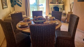 Un coin repas avec des chaises en osier et une table ronde dressée pour le repas, une cheminée et une vue à travers les portes vitrées.