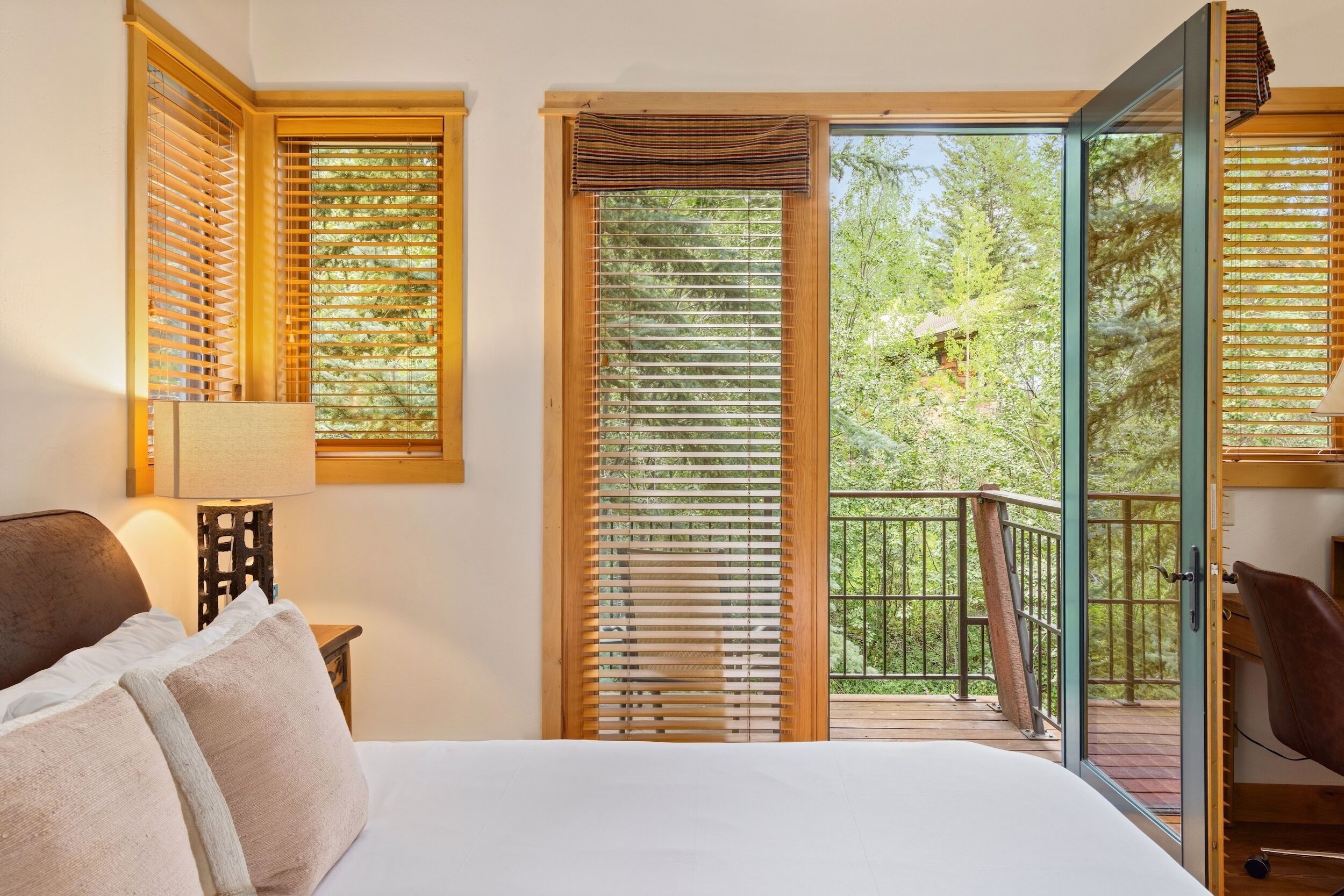 A bedroom with a bed, a nightstand, a lamp, and a view of a balcony with wooden blinds.