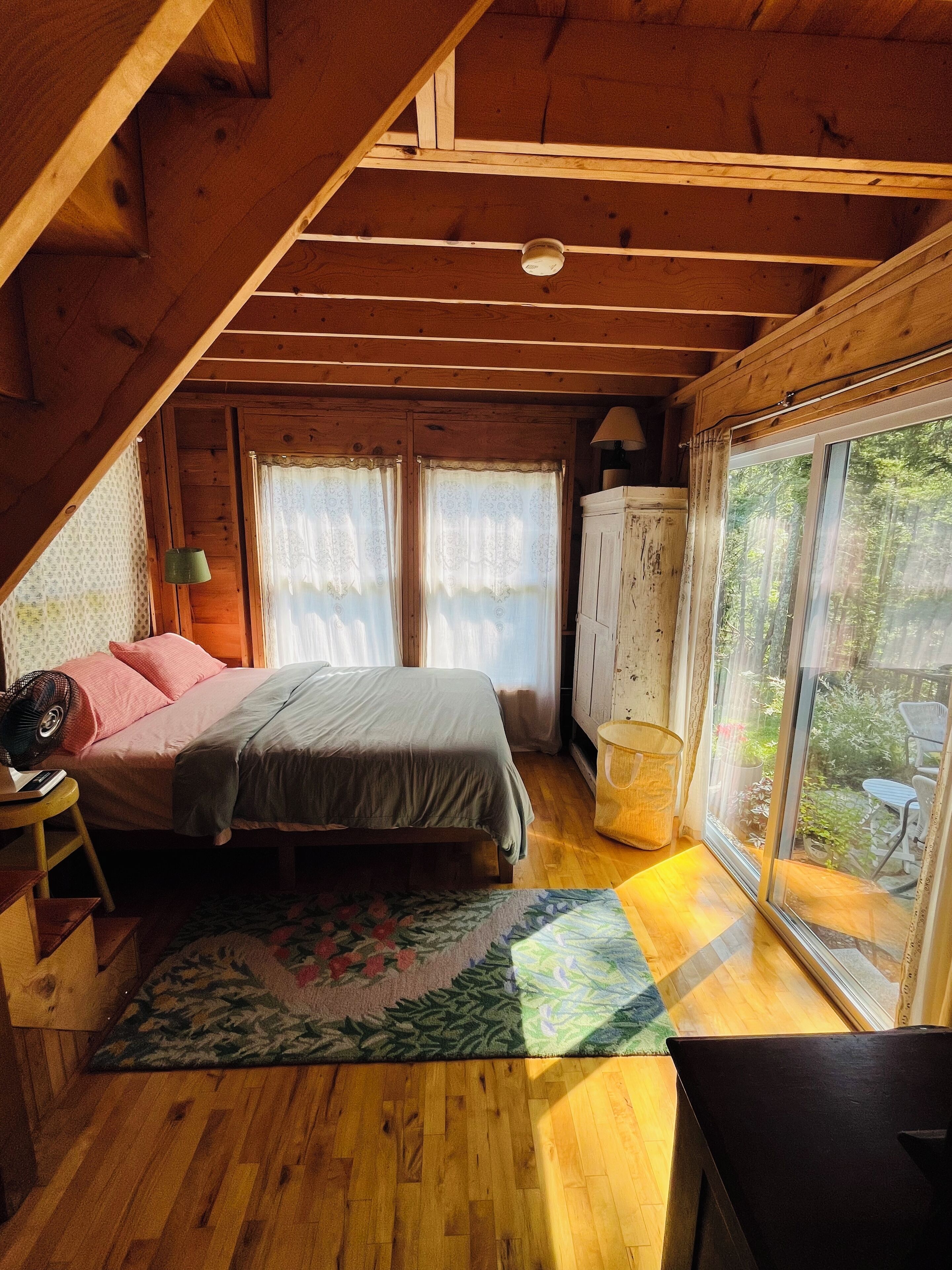 A cozy attic bedroom with a wooden ceiling, a bed with a floral bedspread, a small table with a fan, and a view of the outdoors through a large window.