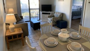 A living room with a sofa, a coffee table, a TV, a dining table with bowls and plates, and a balcony with a view. - Ocean View Unit 3305 - Ocean & Racquet Resort [20% DISCOUNT ON 7+ NIGHT STAYS] (St. Augustine Beach)