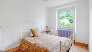 Une chambre avec un lit en métal blanc, une couvre-lit à motifs, une fenêtre donnant sur un paysage verdoyant et une petite table sur laquelle se trouvent un panier et une lampe.