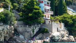 Un bâtiment situé sur la côte, doté d’un escalier en pierre menant à un balcon, entouré de verdure et offrant une vue sur une étendue d’eau.