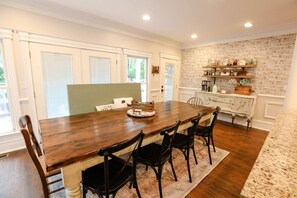 A dining area with a wooden table and black chairs, a brick wall, and a kitchen with white cabinets and a shelf with kitchen items. - Entire Home l Chick Inn on Main (Summerville)