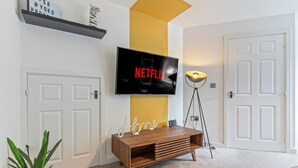 Een moderne woonkamer met een flatscreen televisie aan een geel accentmuur, een houten tv-meubel, een vloerlamp en een potplant.