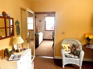 A room with a bed, a chair with floral cushions, a dresser with a lamp, and a door leading to another room.