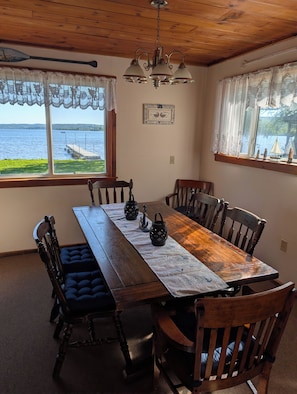 A wooden dining table with a placemat, surrounded by chairs, in a room with a view of a lake and dock.