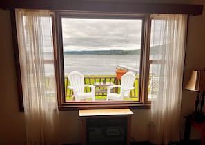 A room with a large window overlooking a body of water, two white chairs, a small table, and a lamp.