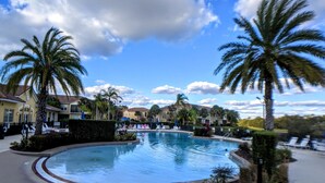 A swimming pool surrounded by palm trees and lounge chairs, with a clear sky and buildings in the background. - Lake View 3BR Condo ...Near Disney and Shopping (Kissimmee)
