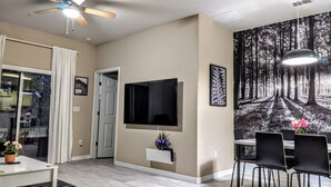 A modern living room with a flat-screen TV, a ceiling fan, a dining area with chairs, and a large forest-themed wall mural. - Lake View 3BR Condo ...Near Disney and Shopping (Kissimmee)