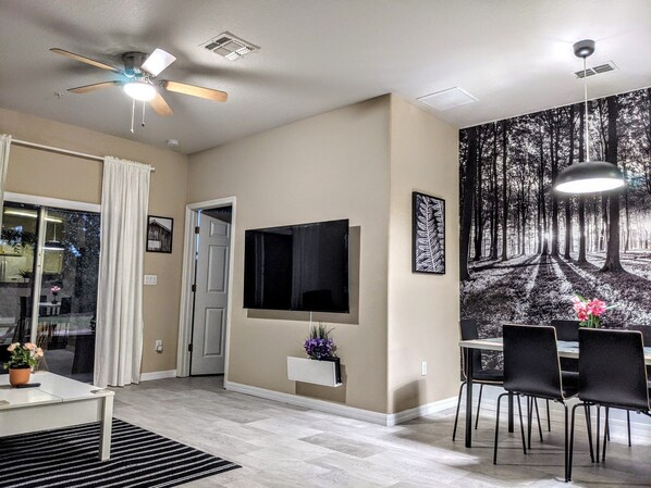Un salon moderne doté d’un téléviseur à écran plat, d’un ventilateur de plafond, d’un coin repas avec des chaises et d’une grande fresque murale sur le thème de la forêt.