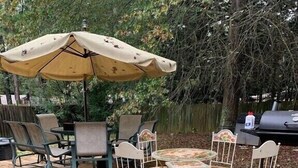 Eine Holzterrasse mit einem runden Tisch, Stühlen, einem Sonnenschirm und einem Grill.
