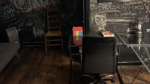 Ein Zimmer mit einer Tafel, die mit verschiedenen Schriftzügen und Zeichnungen bedeckt ist, einem Schreibtisch mit einer Lampe, einem Stuhl und einem kleinen Tisch mit einer bunten Schachtel.