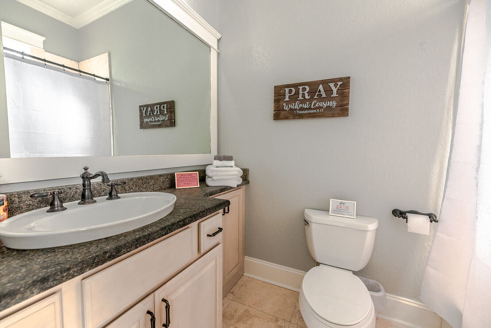 Een badkamer met een wastafel, spiegel en toilet. Op de muur staan borden met de tekst 'PRAY' en 'PRAY Without Ceasing'.