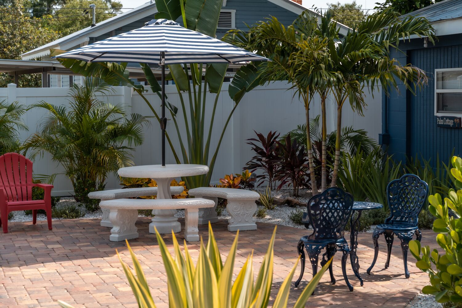 A patio with a striped umbrella, a red Adirondack chair, a white table with a pedestal, and blue chairs.