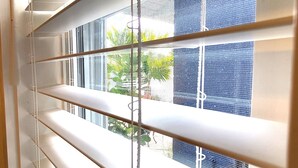 Une fenêtre avec des stores horizontaux entrouverts, laissant entrevoir la verdure à l’extérieur.