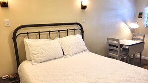 Un lit bien fait, recouvert d’une courtepointe blanche matelassée et agrémenté de deux oreillers, une petite table de chevet avec une lampe, et une chaise placée devant un bureau également équipé d’une lampe.