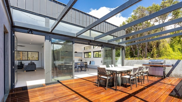 Una casa moderna con terraza de madera, comedor y cocina visibles a través de grandes puertas de cristal.