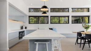 Una cocina moderna con una isla grande, muebles blancos y varias ventanas con vistas a la vegetación.