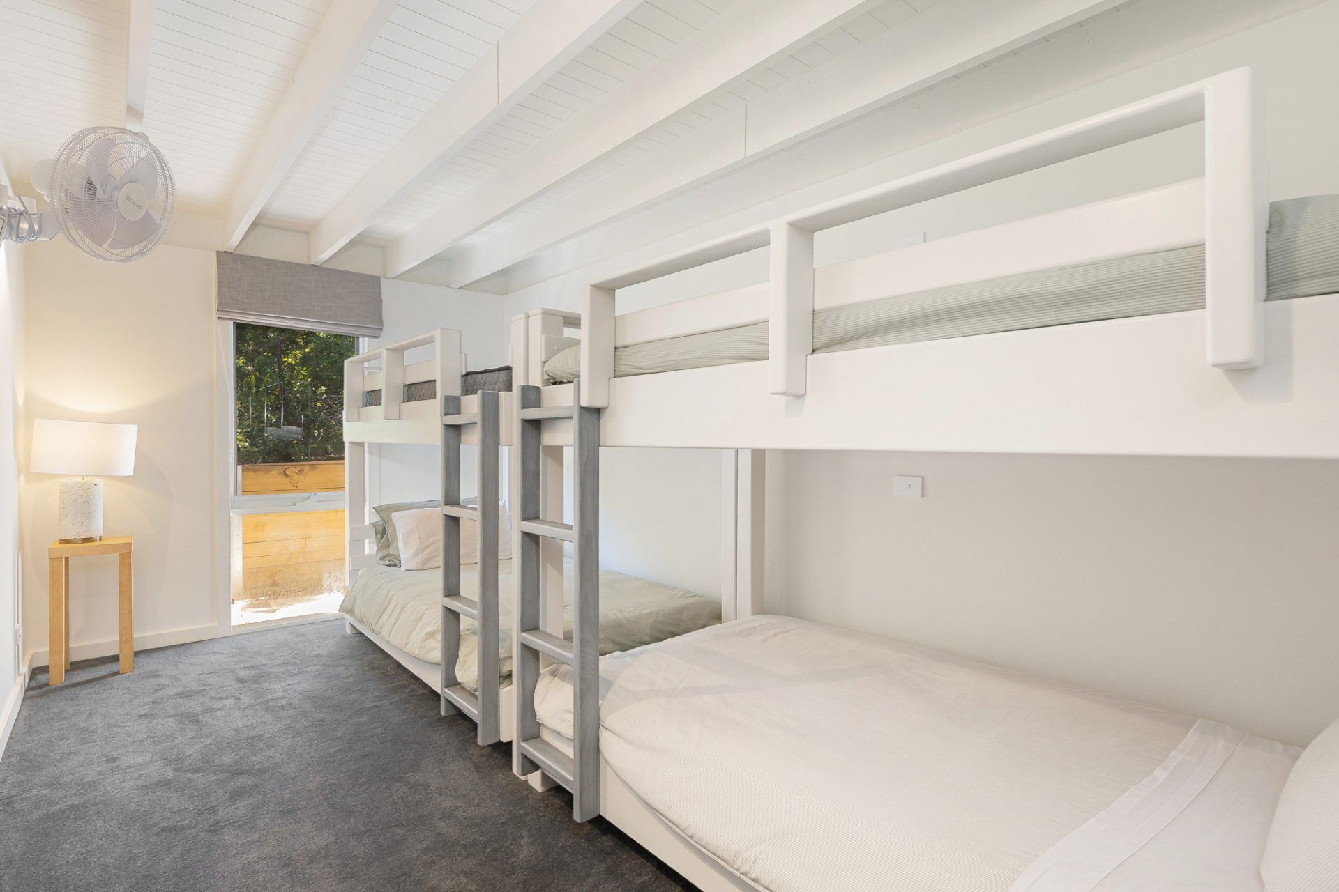 A bunk bed room with a wooden ladder, a small table with a lamp, and a view of greenery outside.