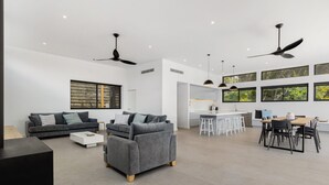 Una sala moderna con un sofá gris, una mesita de centro y un comedor con mesa y sillas. Hay ventiladores de techo, ventanas grandes y una cocina al fondo.