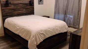 A bedroom with a wooden bed frame, a white bedspread, a nightstand, a lamp, and a window with a curtain.