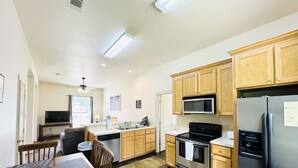 A modern kitchen with wooden cabinets, stainless steel appliances, a dining area with a table and chairs, and a television in the background. - Chateau 11, spacious townhouse in Tickfaw, LA (Tickfaw)