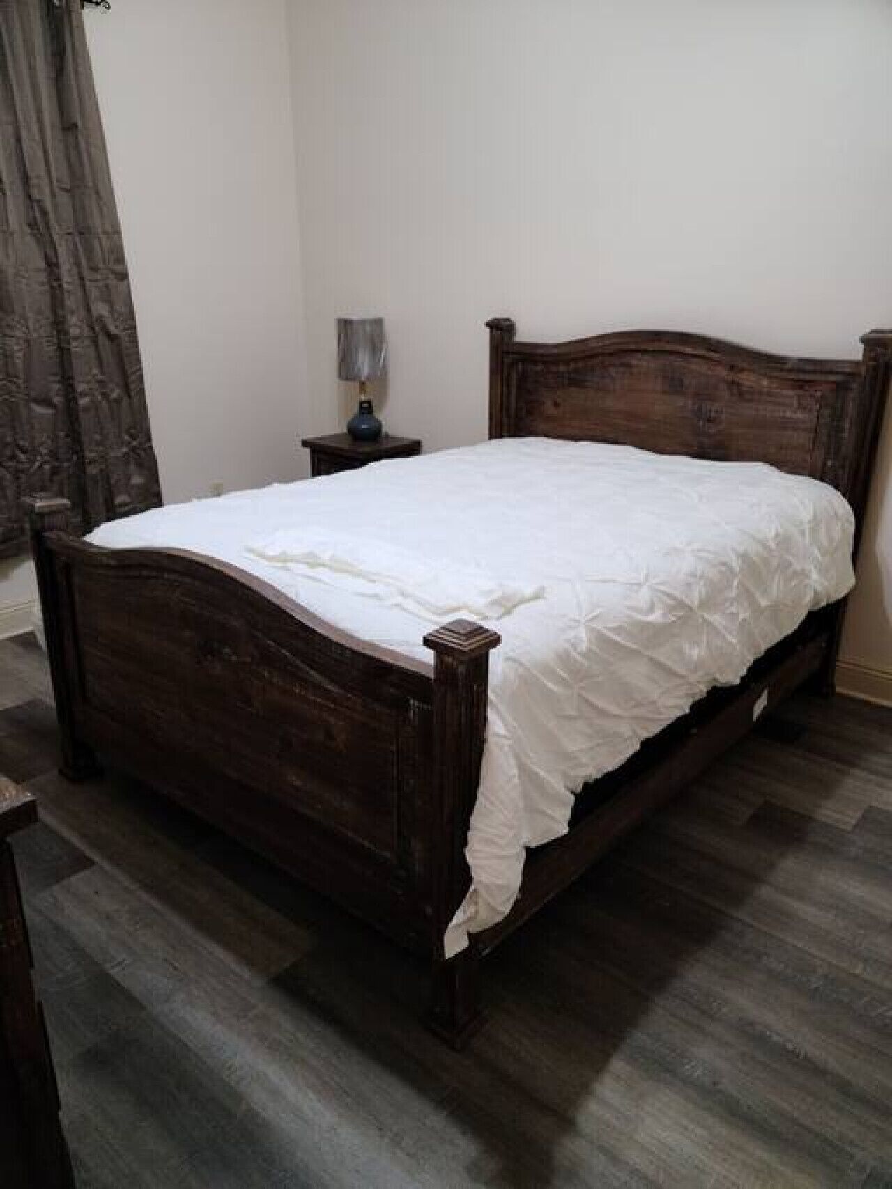 A wooden bed with a white bedspread in a room with a dark wooden floor and a nightstand with a lamp.