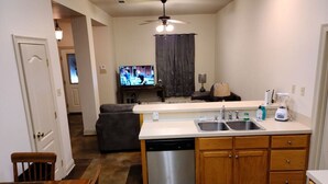 A compact kitchen with a dishwasher, a sink, and wooden cabinets. There is a dining area with a table and chairs, and a television in the background. - Chateau 11, spacious townhouse in Tickfaw, LA (Tickfaw)
