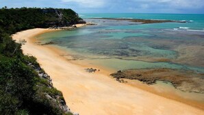 A beach with clear turquoise waters, sandy shores, and a lush green cliff. - Estúdio Martinelli (Porto Seguro)
