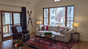 A spacious living room with a large window, a leather armchair, a white sofa with red cushions, a coffee table, and a red patterned rug. - Downtown Loft on Lupfer (Whitefish)