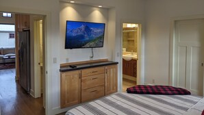 A bedroom with a bed, a TV mounted on the wall, wooden cabinets, and a bathroom visible through an open door. - Downtown Loft on Lupfer (Whitefish)