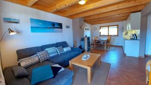 A cozy living area with a sofa, coffee table, and a painting of a beach scene. A kitchen area is visible in the background with a dining table and chairs. - Beautiful, cozy and comfortable vacation home on Lake Lechsee right on the edge of the forest (Lechbruck am See)