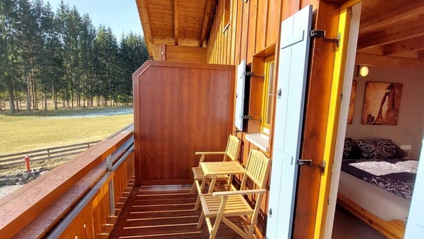 A wooden deck with a view of a forest, a bed, and a door leading to a bedroom. - Beautiful, cozy and comfortable vacation home on Lake Lechsee right on the edge of the forest (Lechbruck am See)