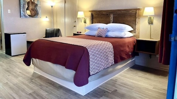 Une chambre d’hôtel avec un grand lit, deux lampes de chevet, un petit bureau avec une lampe, une œuvre d’art accrochée au mur et une porte.