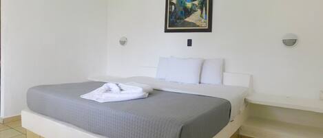 A neatly made bed with white linens and a grey blanket, a painting on the wall, and a small bedside table with a lamp.