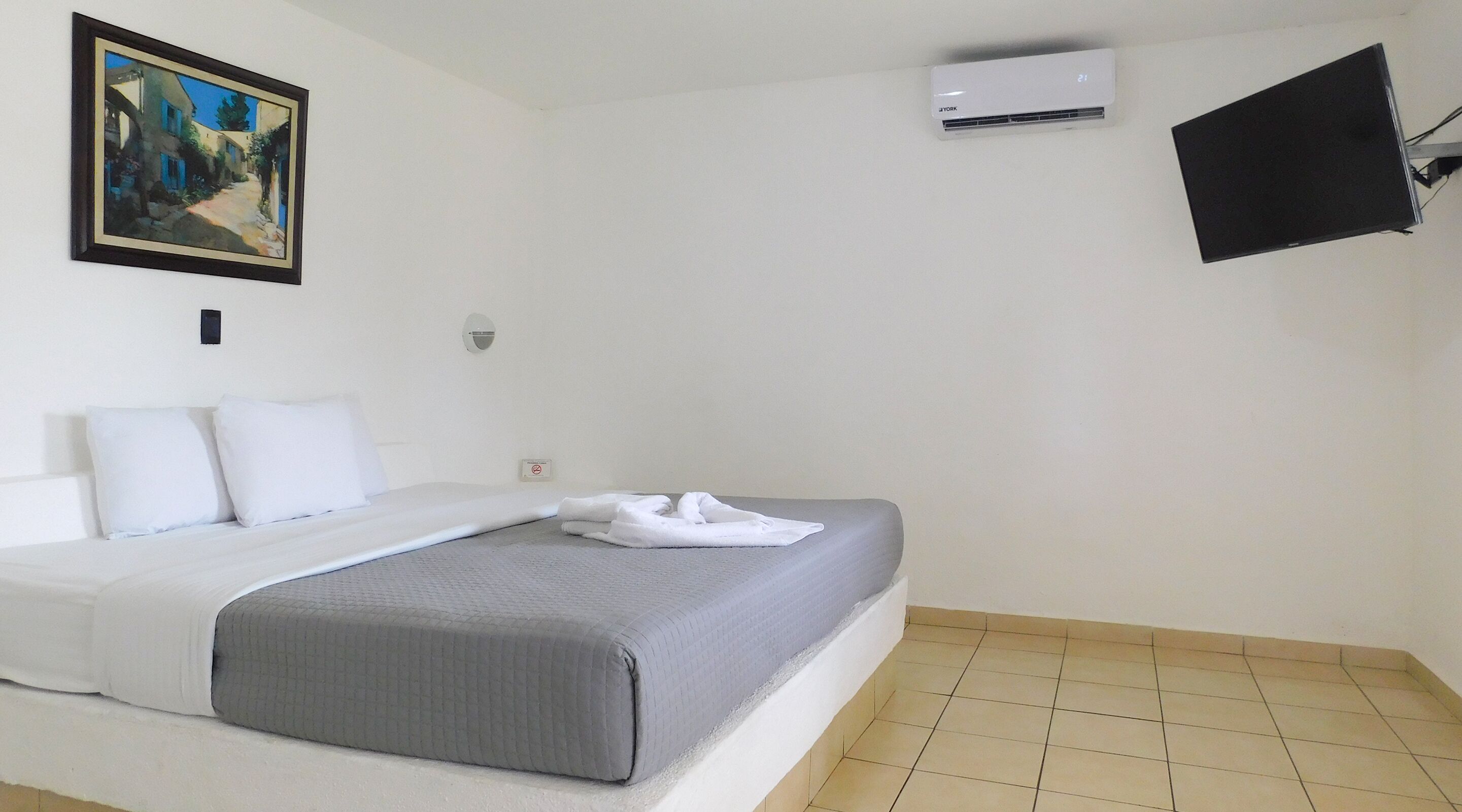 Habitación de hotel con cama, almohadas blancas, manta gris, un cuadro en la pared, aire acondicionado y televisor montado.