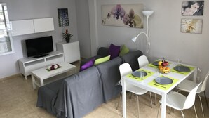 Un salon moderne avec un canapé gris, un téléviseur à écran plat, une table à manger avec des chaises et un petit meuble blanc.